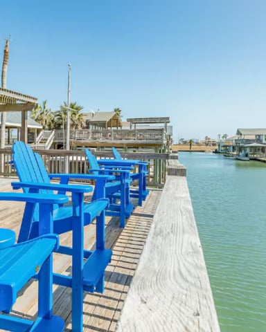 Blue Adirondack chairs on wooden dock overlooking calm waterway with beach houses and palm trees