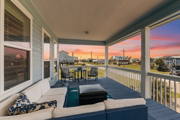 Covered deck with seating area overlooking coastal neighborhood at sunset, orange sky visible beyond houses and utility poles