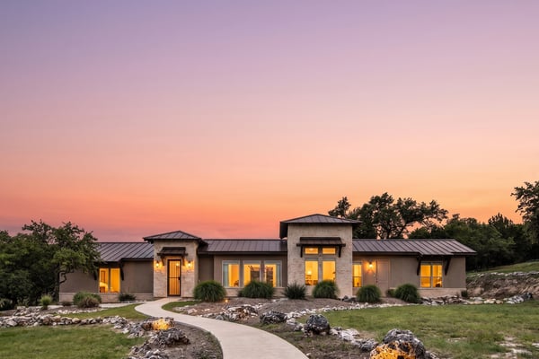 Modern ranch house with metal roof and stone accents at sunset, curved walkway through landscaped grounds with ornamental grasses