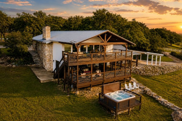 Modern ranch house with elevated deck, hot tub, and stone chimney surrounded by oak trees at sunset