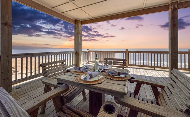 Covered beachfront deck with dining table set for dinner, wooden railings, and sunset ocean view