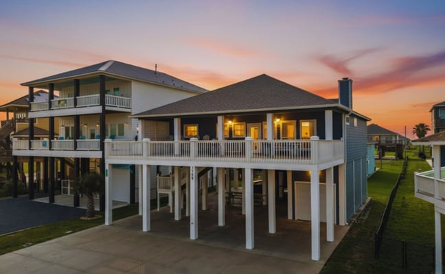 Elevated coastal home with white railings and dark roof at sunset, illuminated windows, concrete driveway, neighboring houses visible.